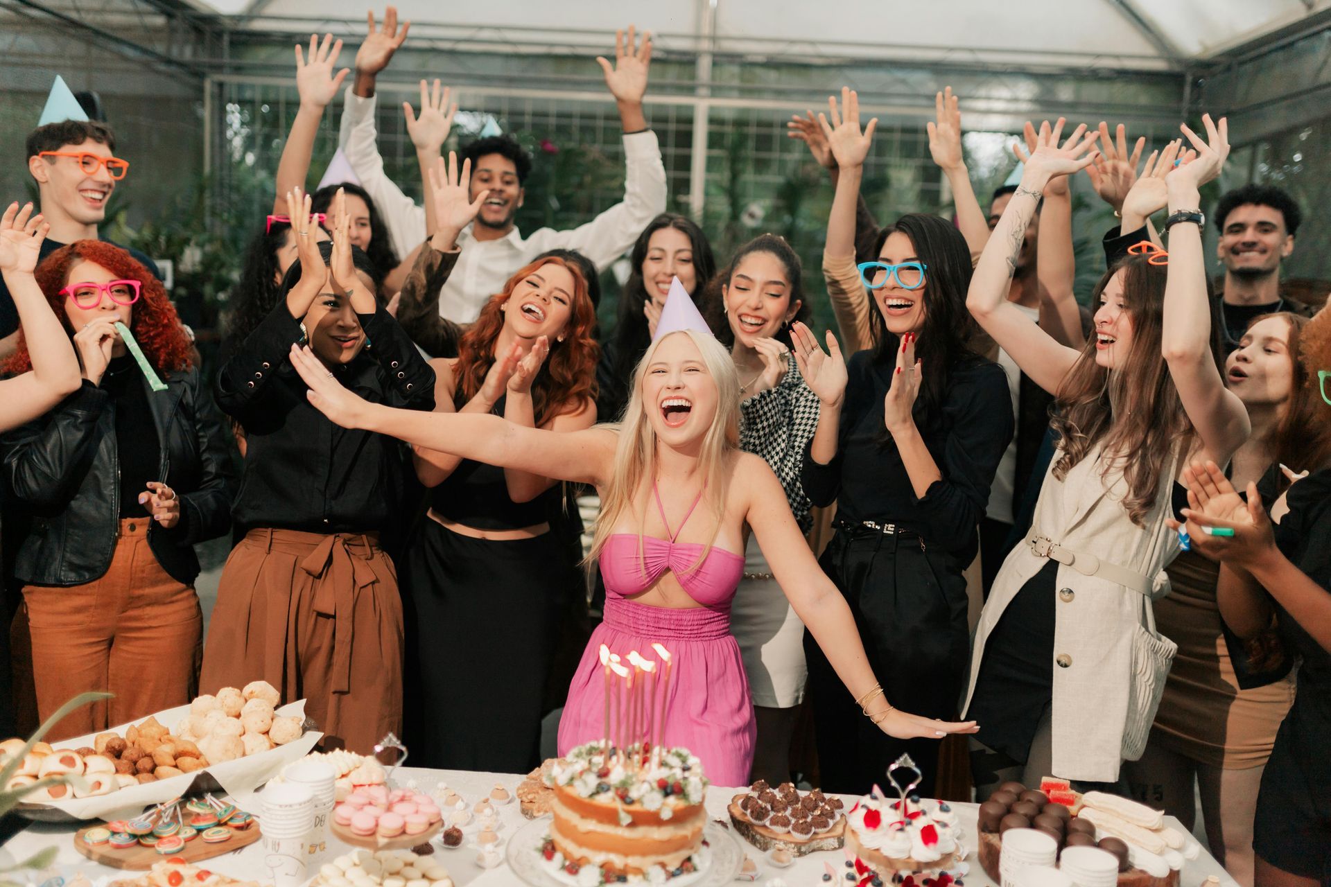 Group of people celebrating a birthday, raising arms, and cheering. A woman in a pink dress stands in front of a cake.