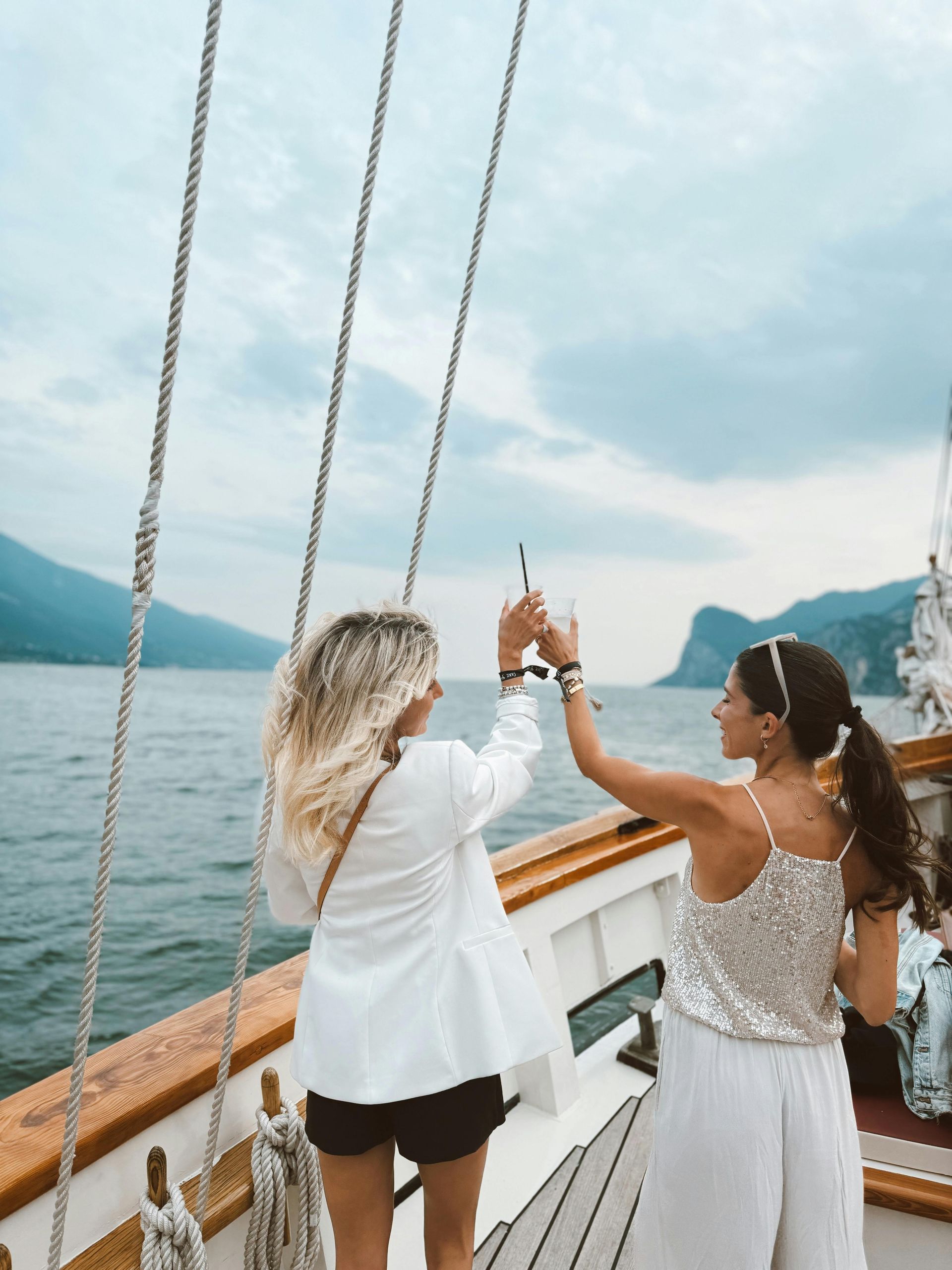 Two women on a boat toast with drinks, against a backdrop of water and mountains.