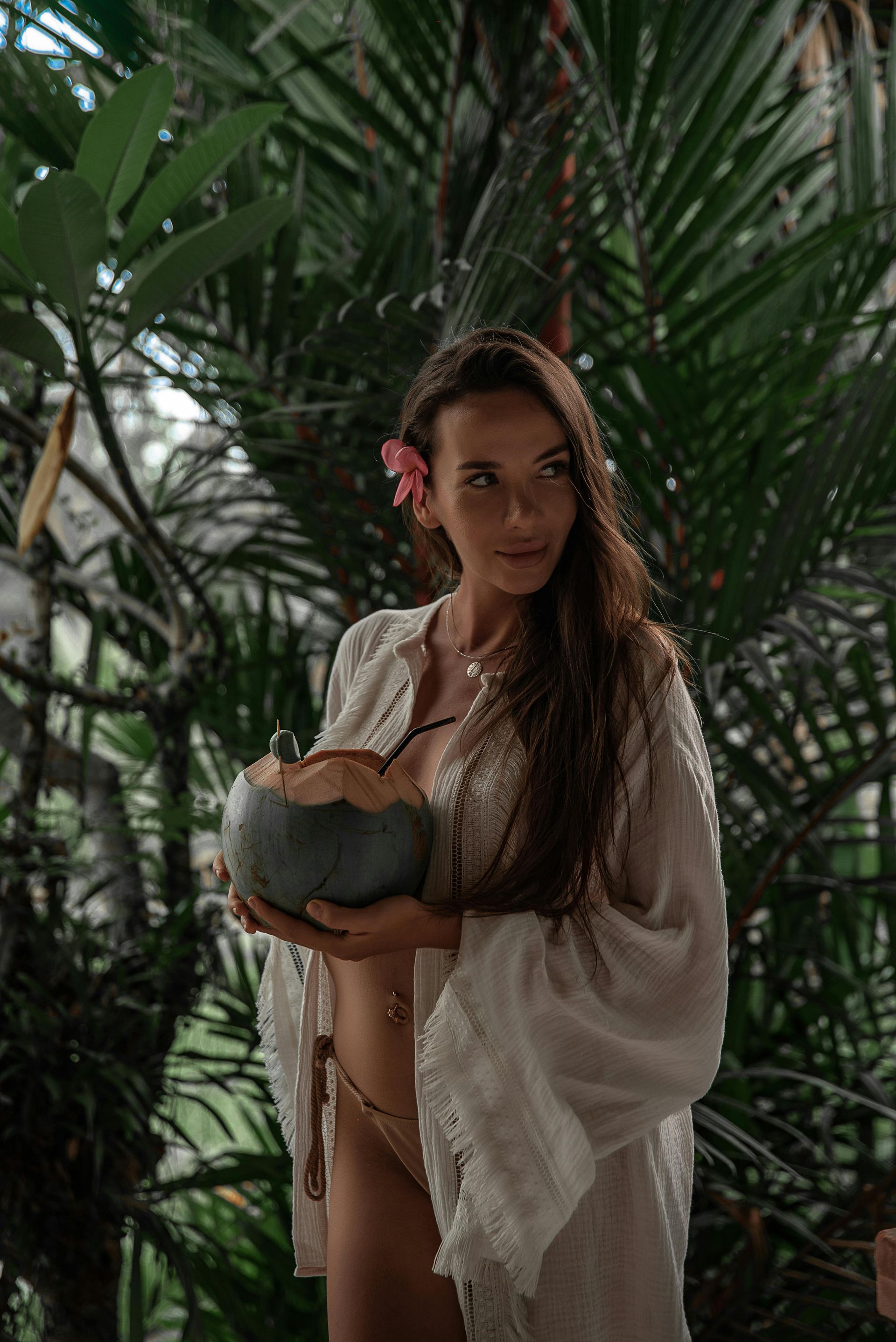 Woman in white robe with a coconut drink, pink flower in hair, surrounded by lush greenery.