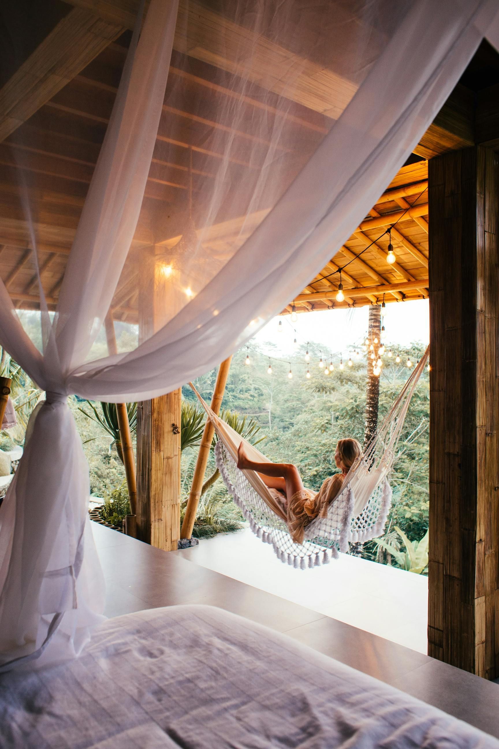 Woman relaxing in hammock on a porch, white mosquito net, wood beams, overlooking lush green foliage.