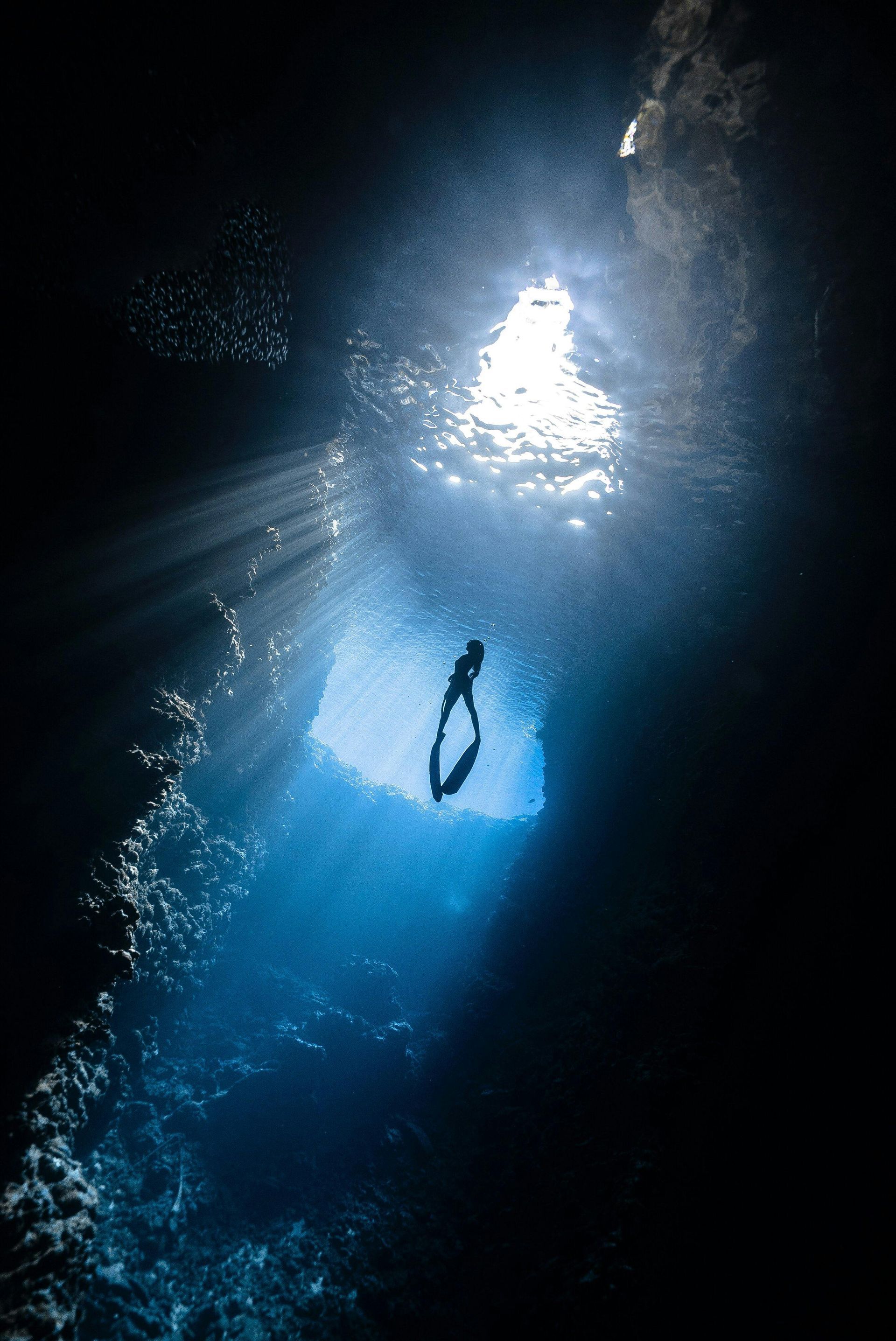 Diver silhouetted in a cavern, sunlight streams down, illuminating the blue water.