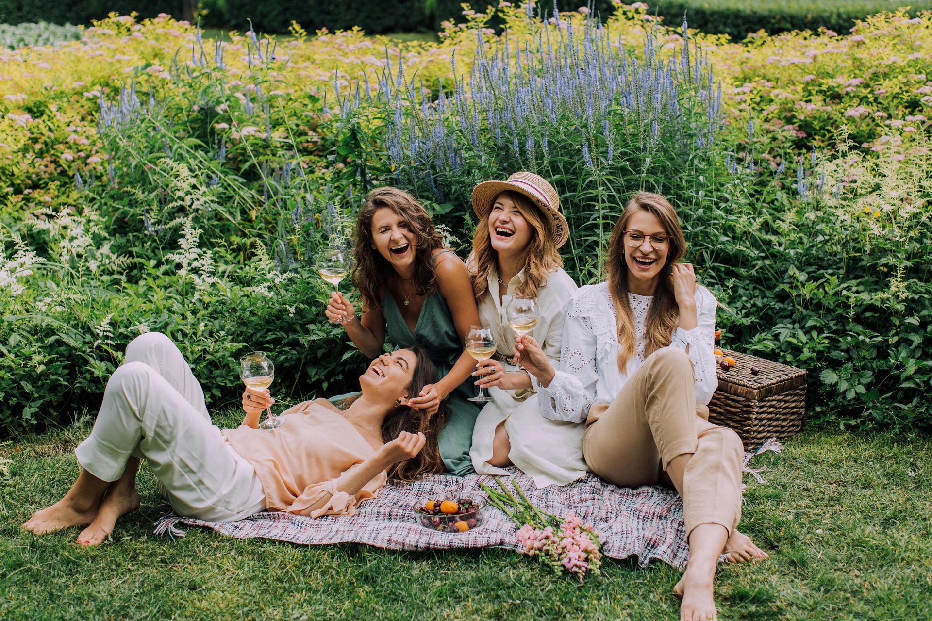 Four laughing women on a picnic in a sunny garden; drinking wine, lying and sitting on a blanket.