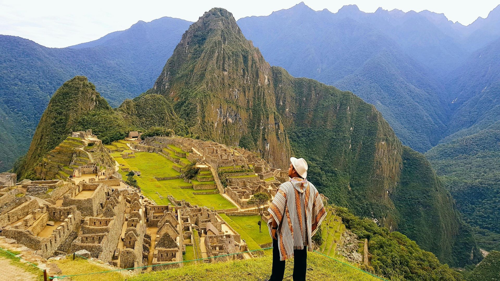 Man in poncho looking at Machu Picchu ruins, mountains in background.