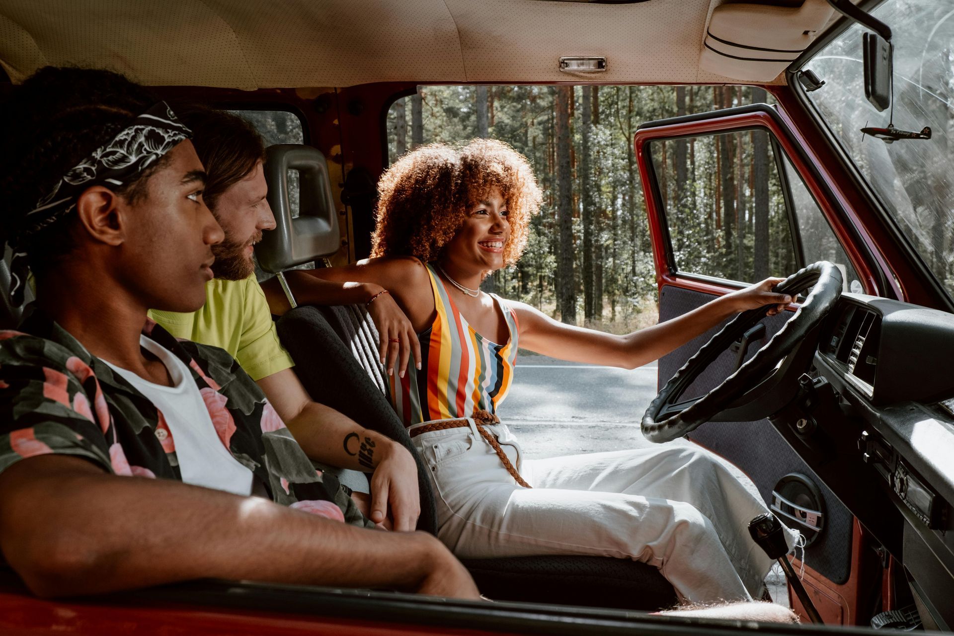 Three friends in a red van, woman driving smiles. Road and trees visible through open door.