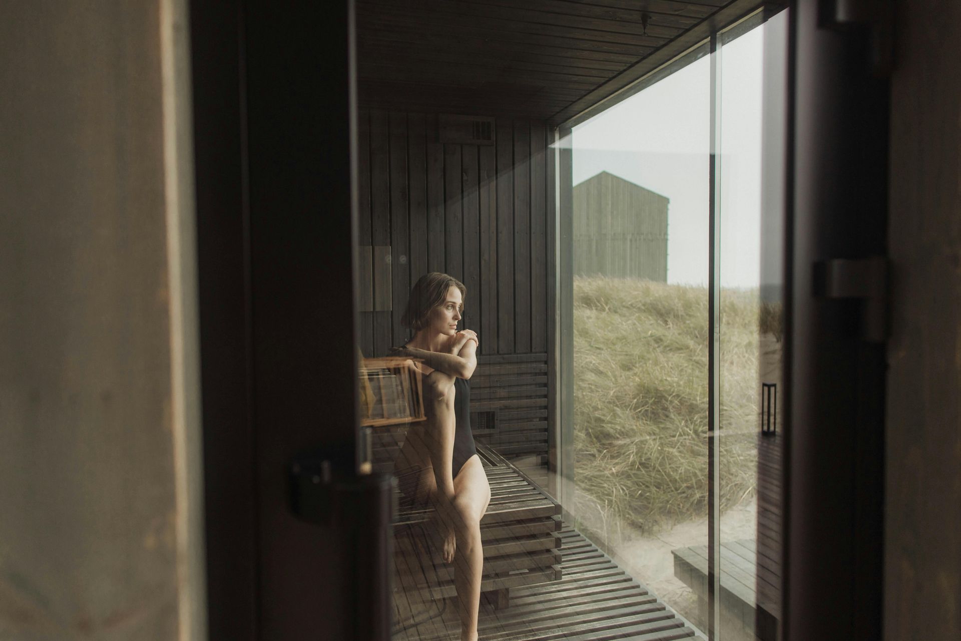 Woman in a black swimsuit sits by a window, looking out at dunes, with wood accents.