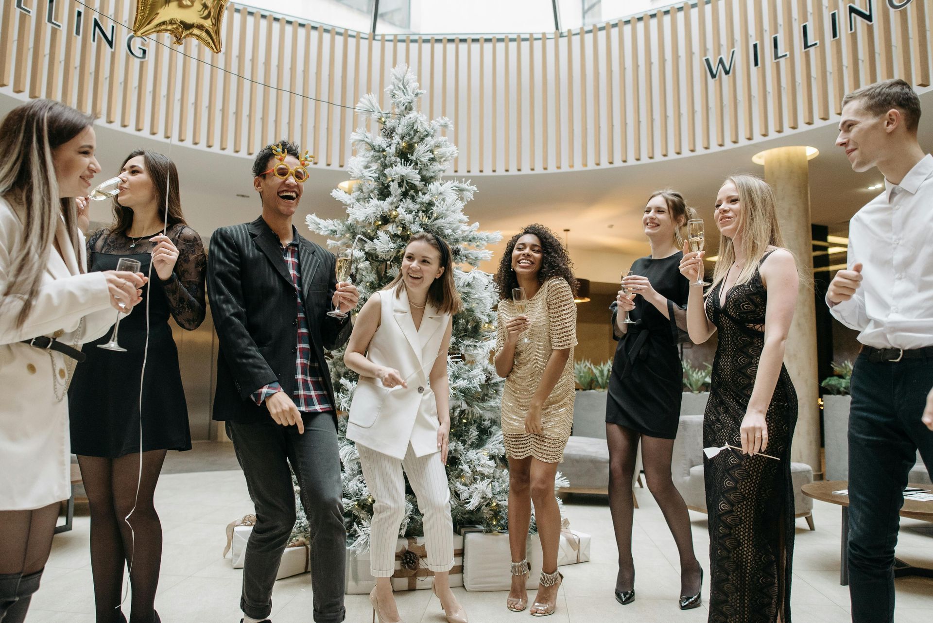 Group of diverse people dancing, celebrating near a Christmas tree, in a modern, well-lit space.