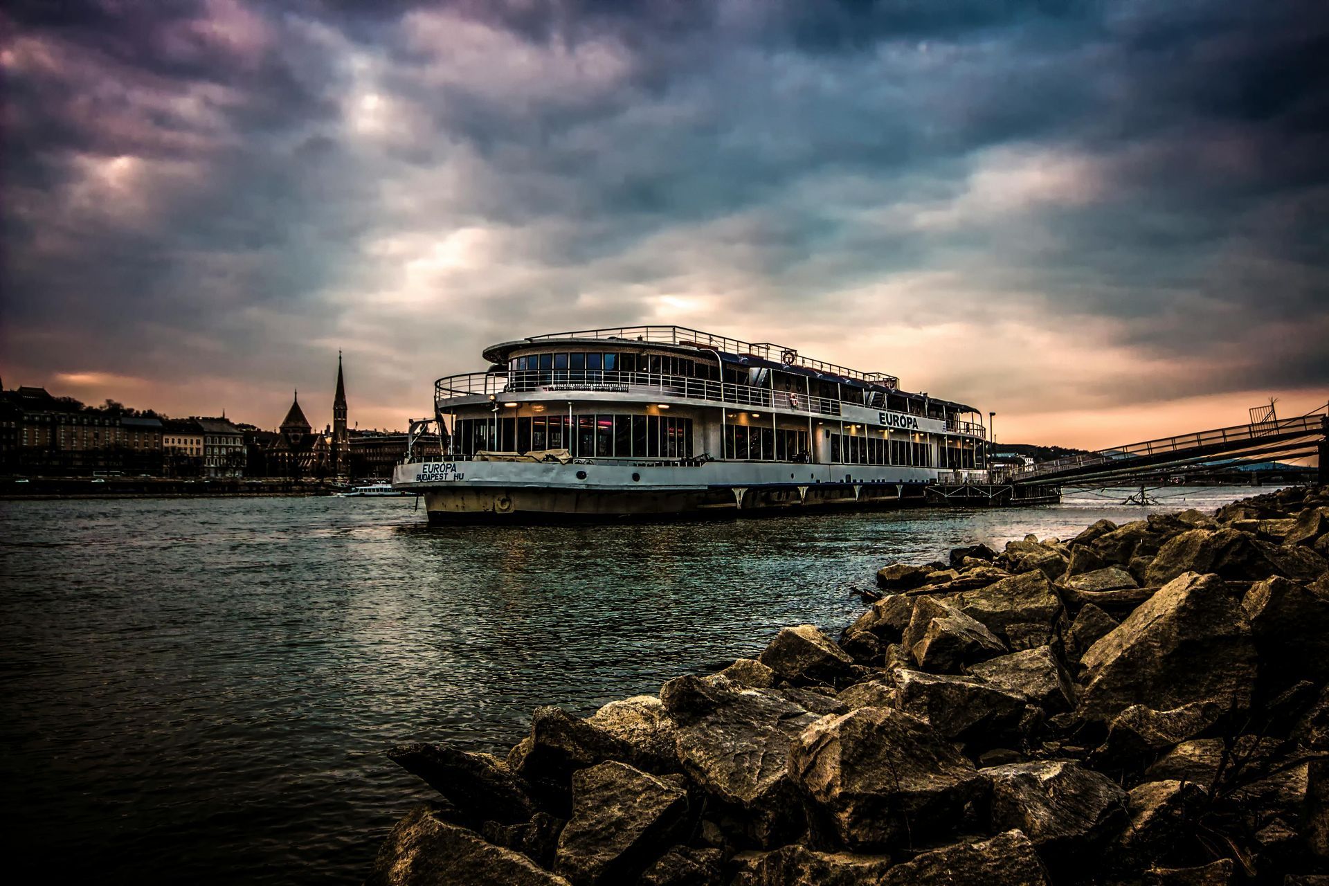 Riverboat on a river with a rocky shoreline, buildings in the background under a cloudy, colorful sky.