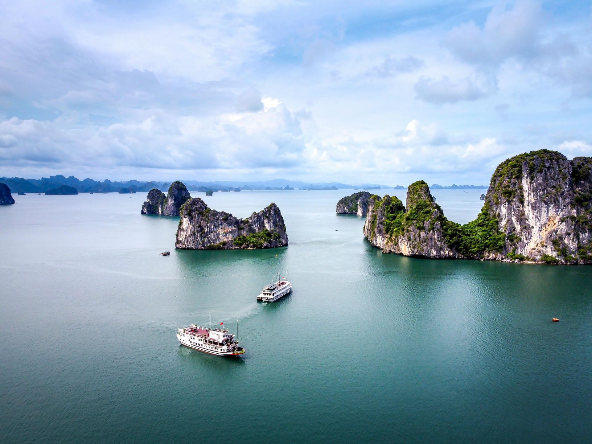 Boats sail through emerald waters dotted with karst islands under a cloudy sky in Ha Long Bay, Vietnam.