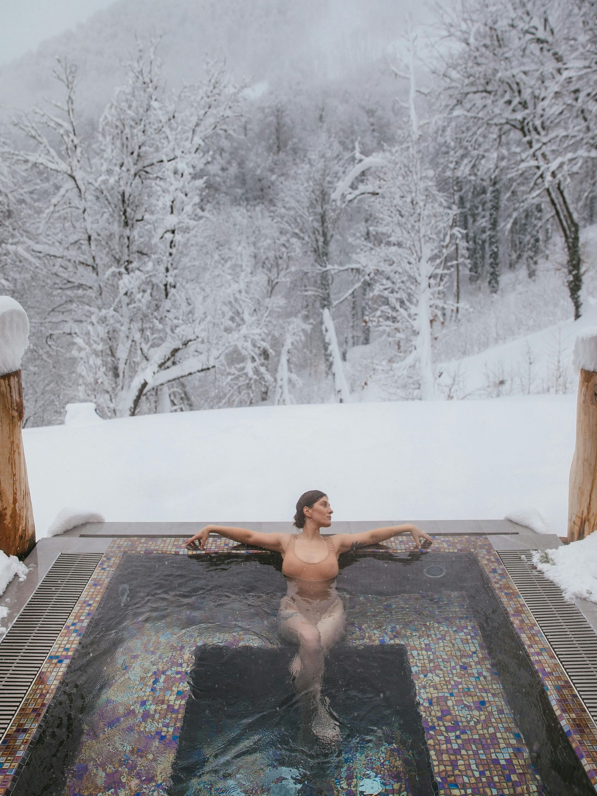 Woman relaxes in outdoor pool, snow-covered trees in background.