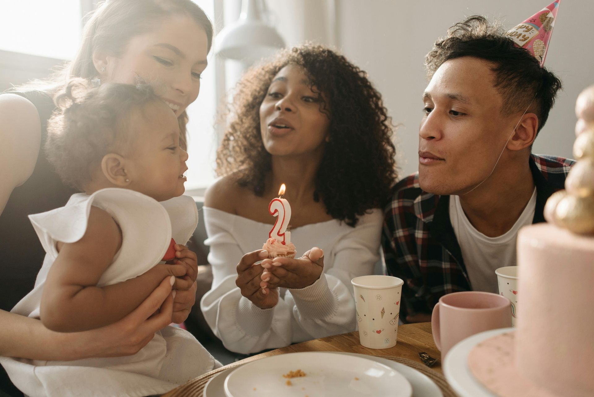 Family celebrates a toddler's 2nd birthday. They gather around a cake, blowing out the candle.