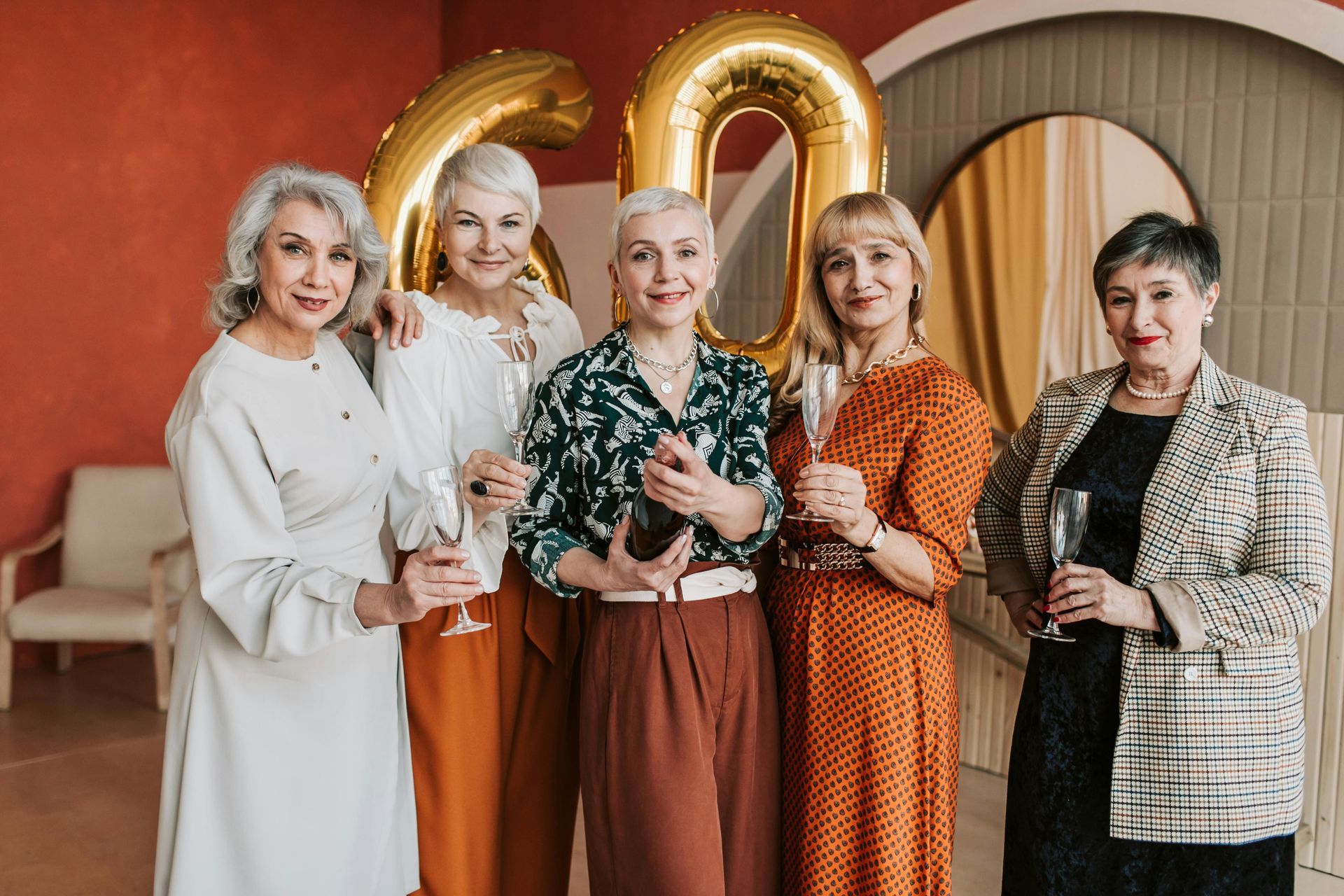 Five women celebrating a 60th birthday. They hold champagne glasses and smile in front of gold balloons.