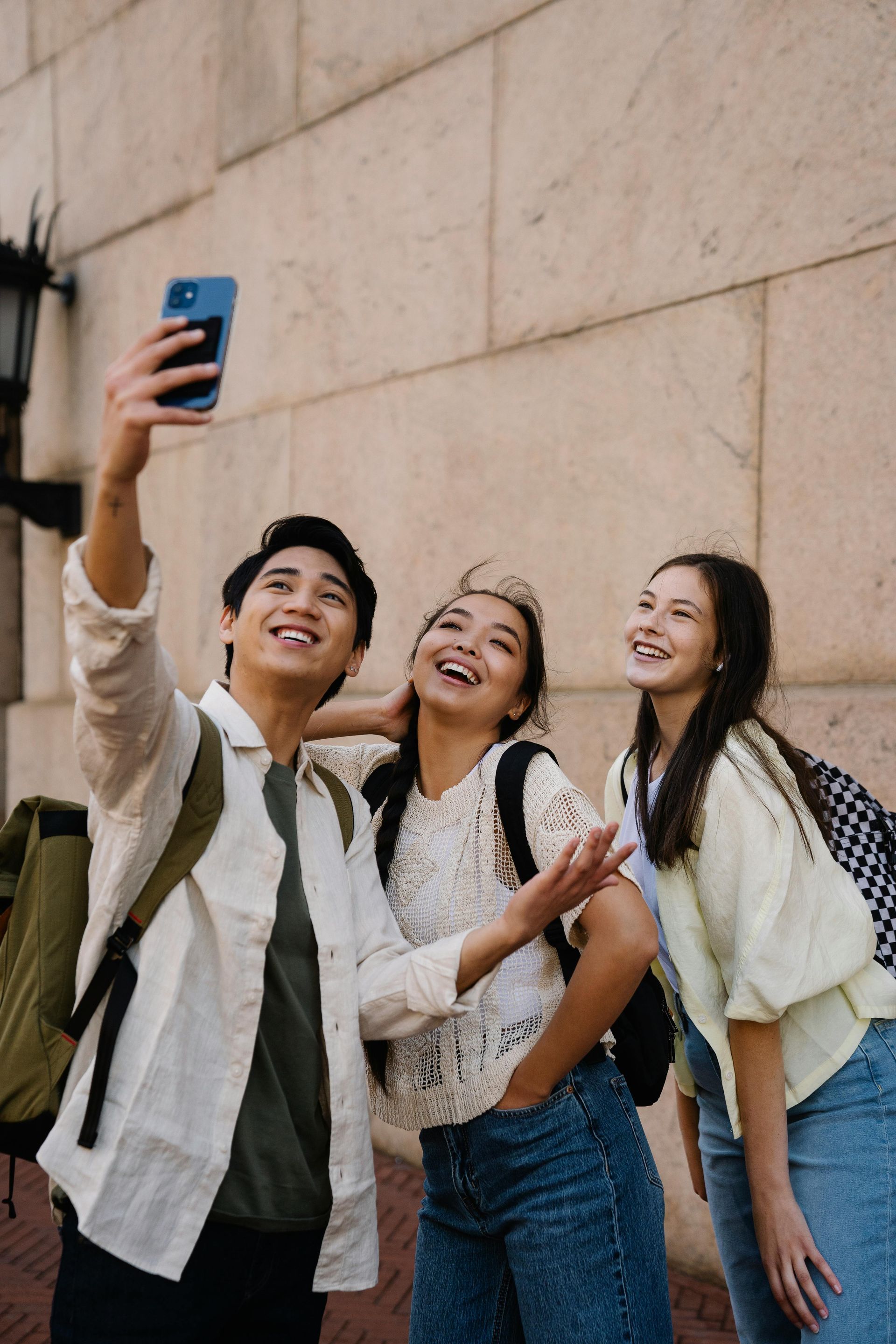 Three young adults smile while taking a selfie against a beige building.