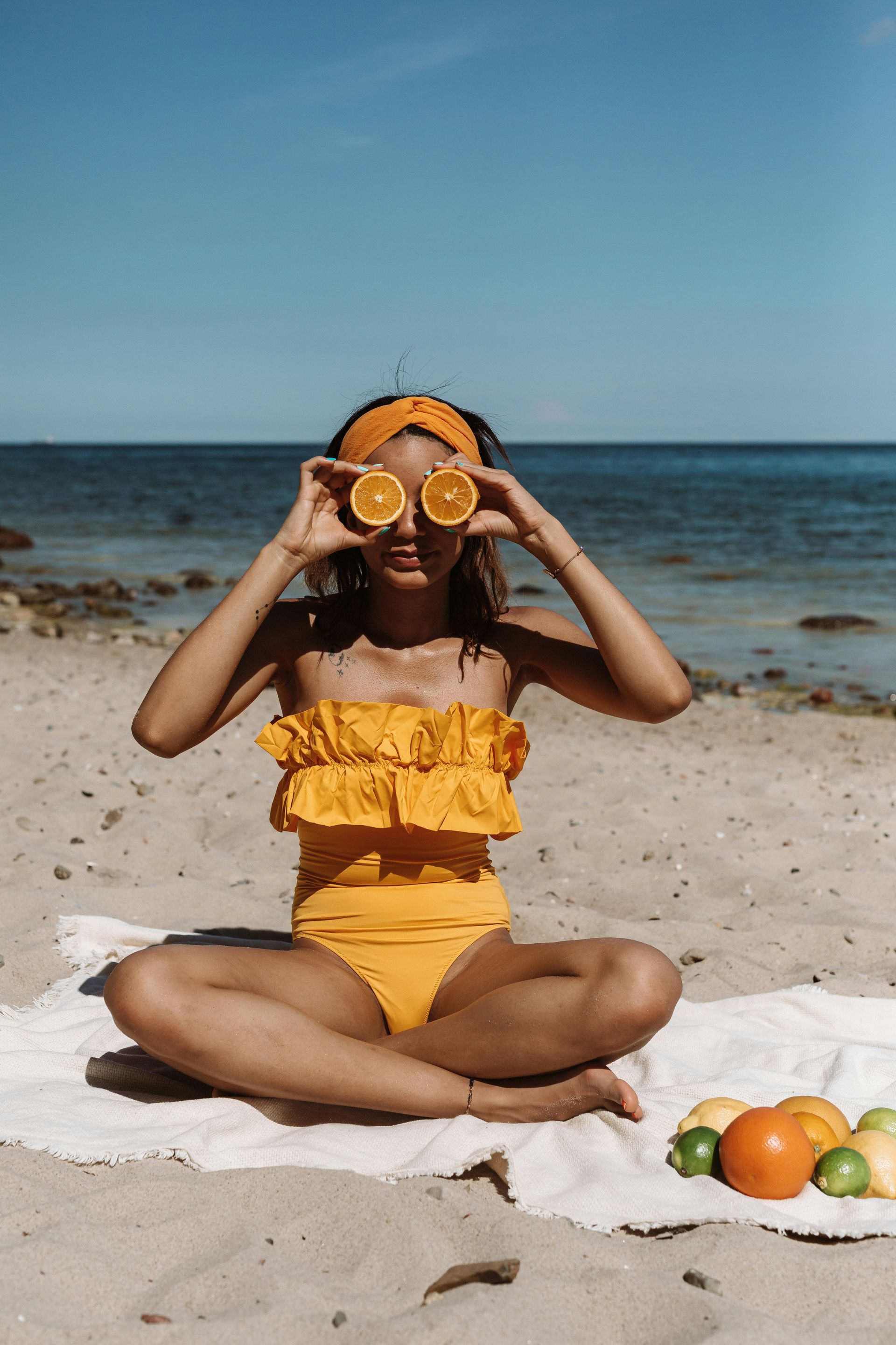 Woman on beach holding orange slices over eyes, wearing yellow swimsuit. Ocean in background.