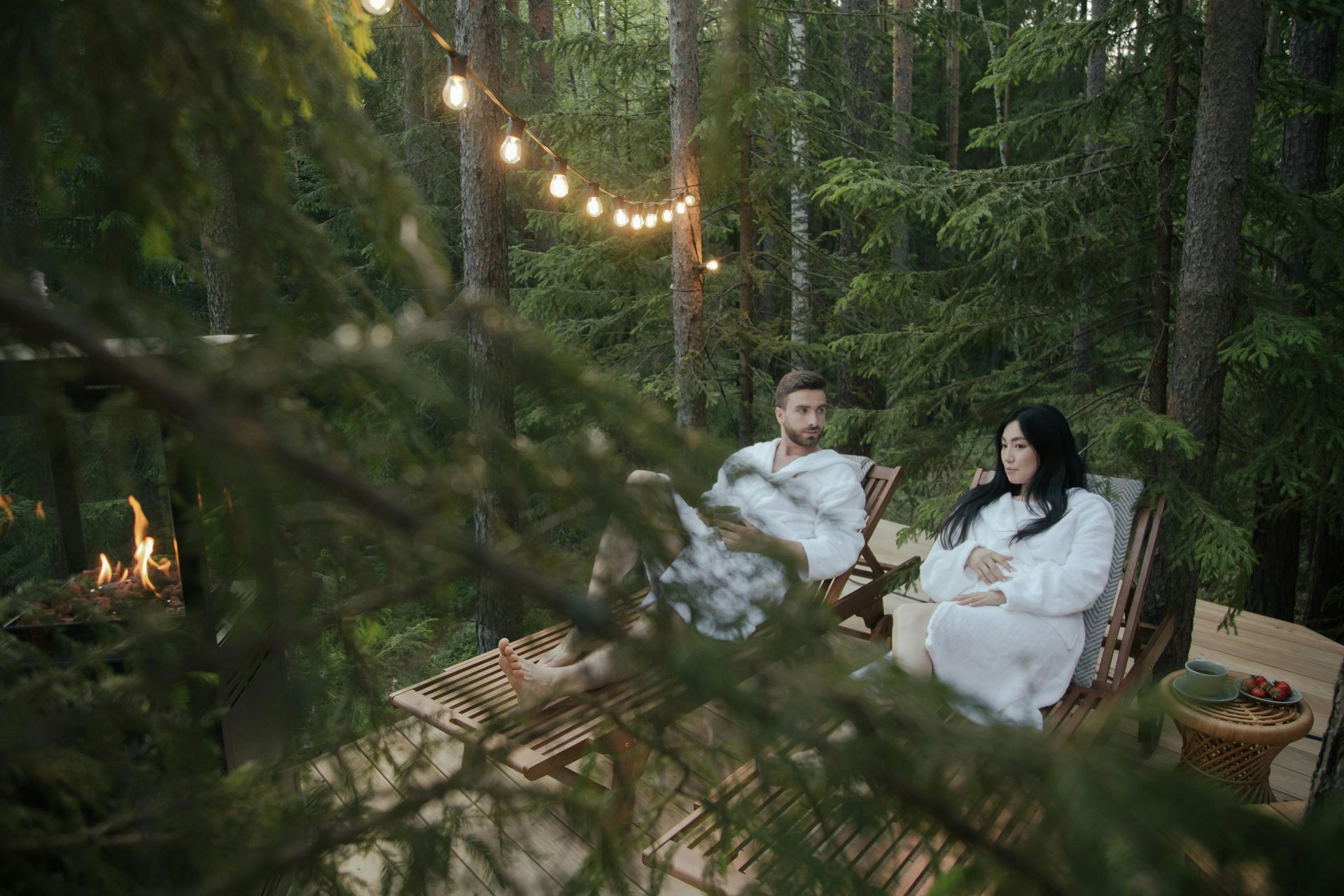 Couple in bathrobes relax in forest chairs under string lights. Fire burns nearby.