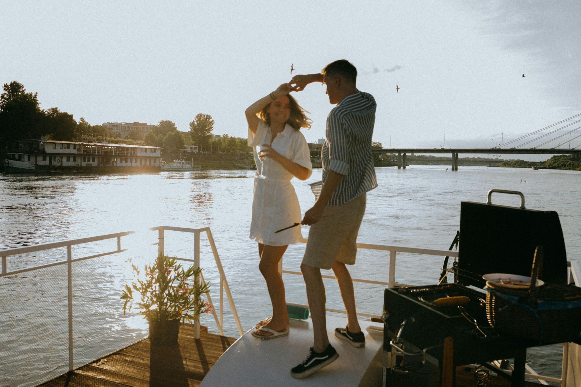 Couple dancing on a boat, grilling in the background, with a bridge and buildings on the river in the sunlight.