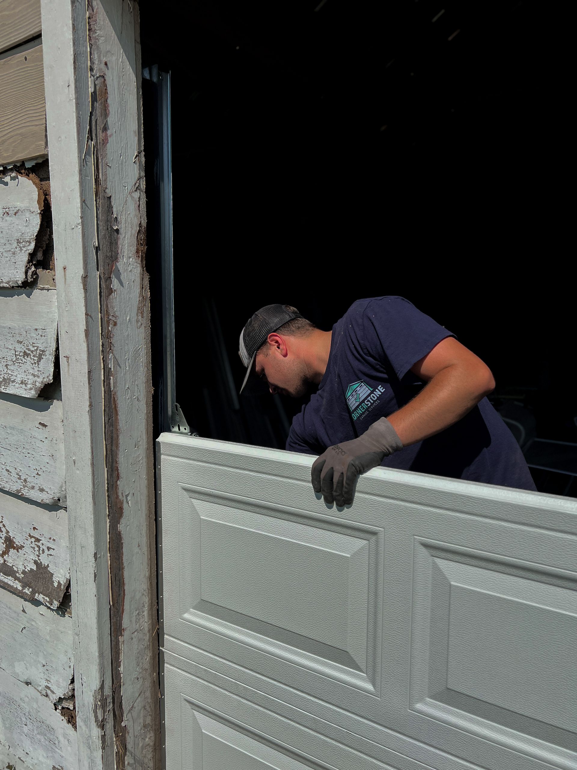 Man installing a garage door panel in a garage doorway. Gray panel, gray gloves. Outdoors, sunny.