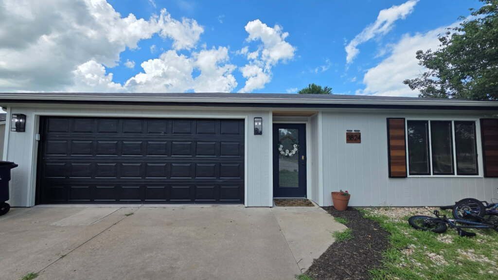Exterior of a light-colored house with a black garage door and door trim; blue sky with clouds.