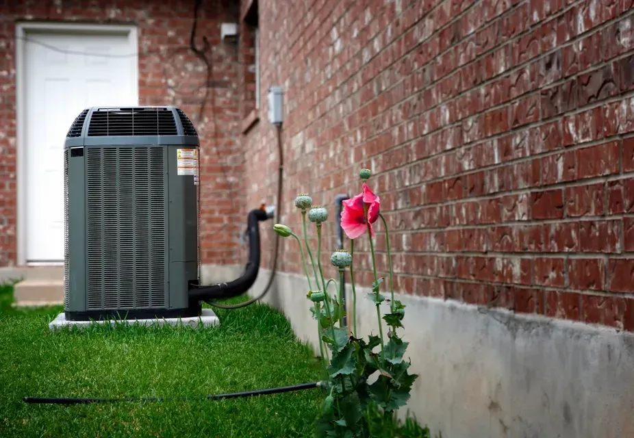 An air conditioner is sitting on the side of a brick building next to a flower.