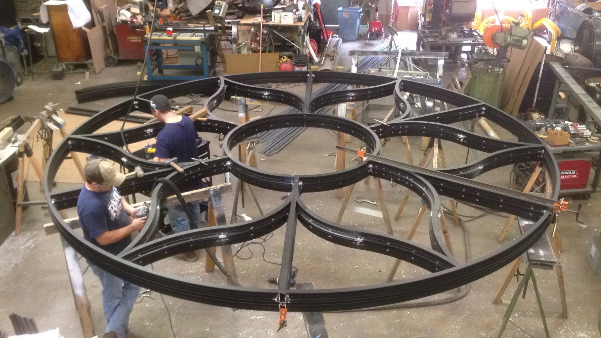 Two men are working on a large metal circle in a workshop