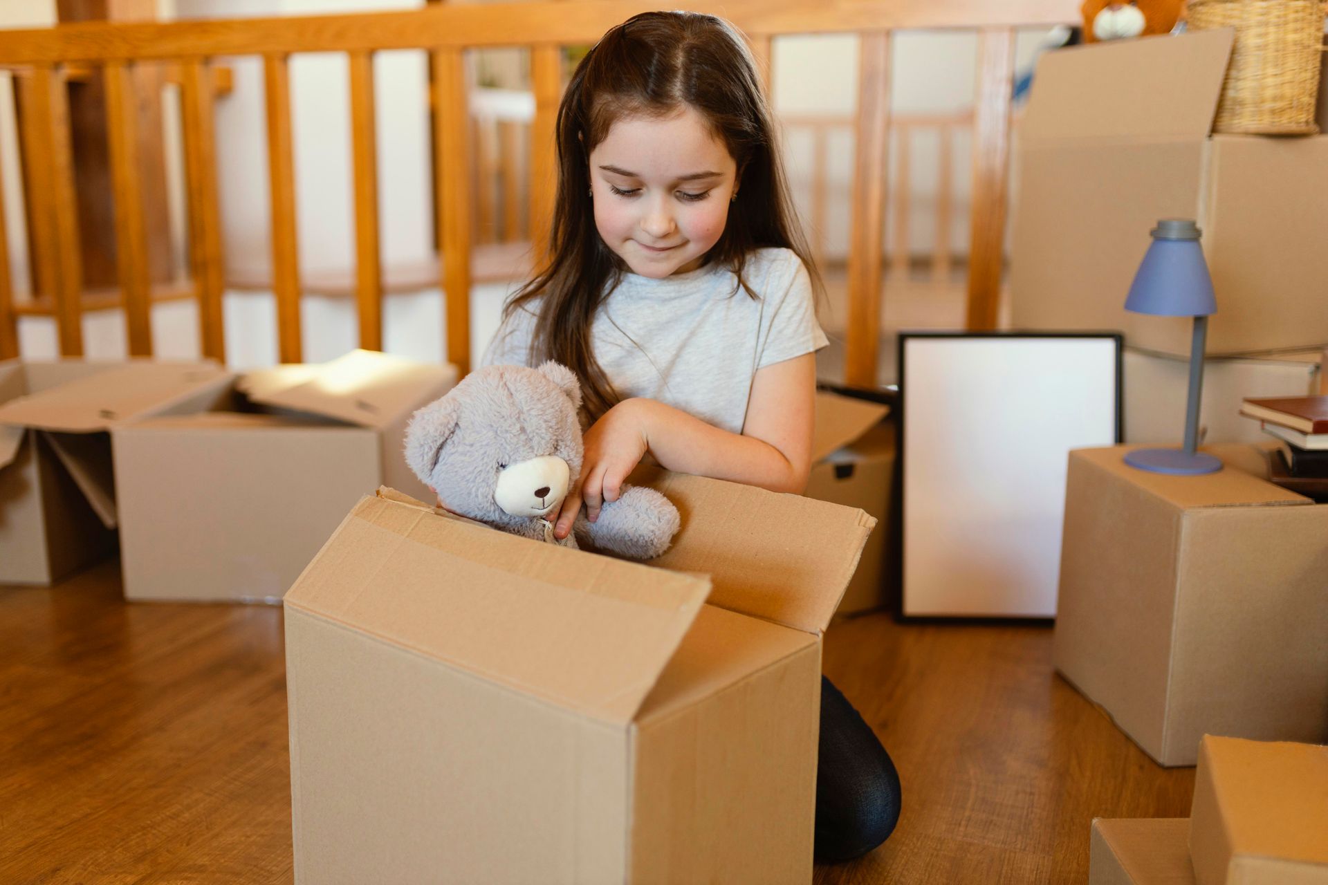 A little girl is sitting on the floor holding a teddy bear in a cardboard box.