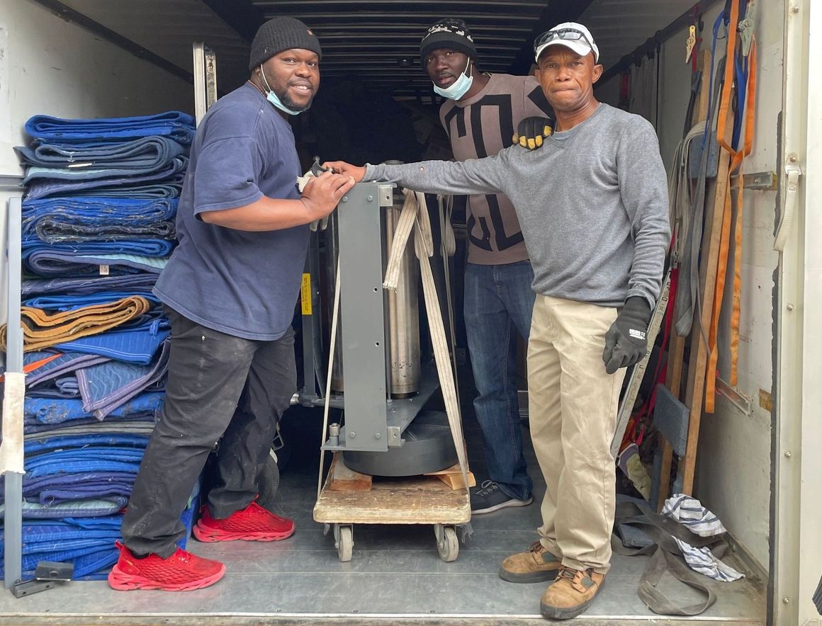 Three men inside a moving truck, loading a large appliance onto a dolly. The men are smiling and wearing work clothes.