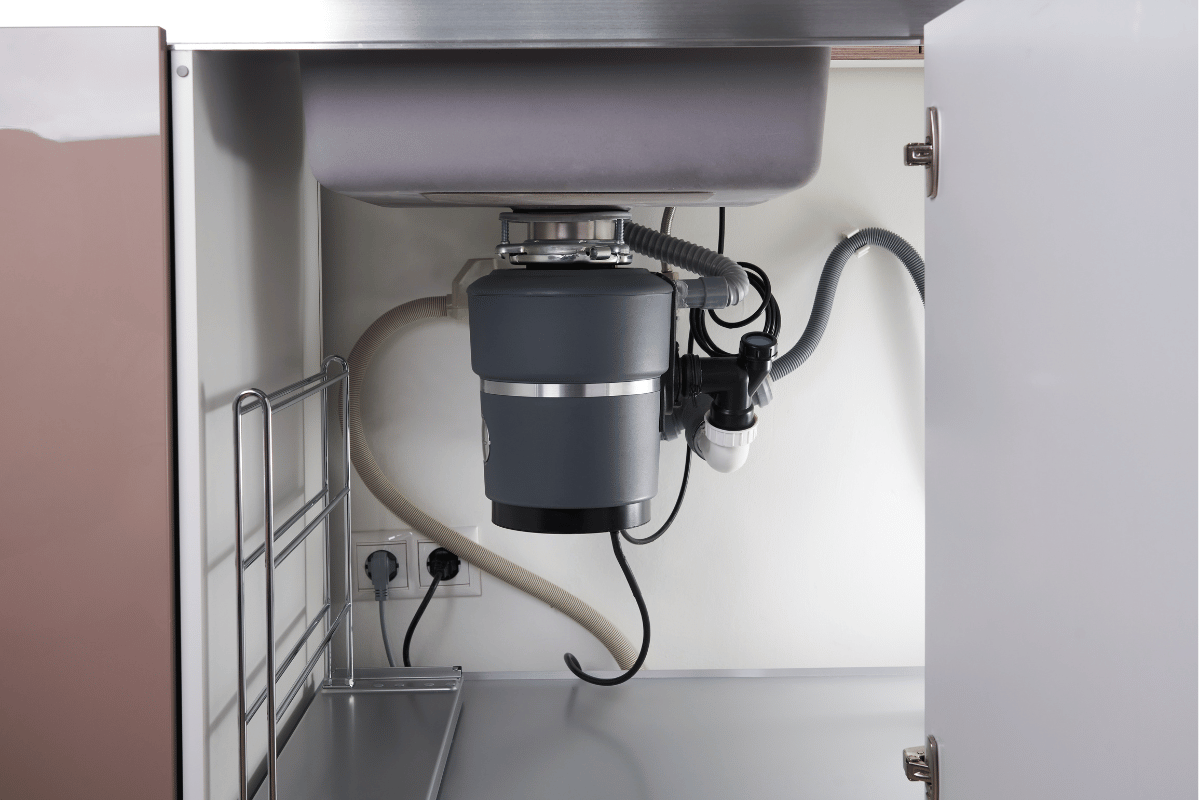 Inside a kitchen cabinet: garbage disposal beneath a sink. Plumbing and electrical components visible.