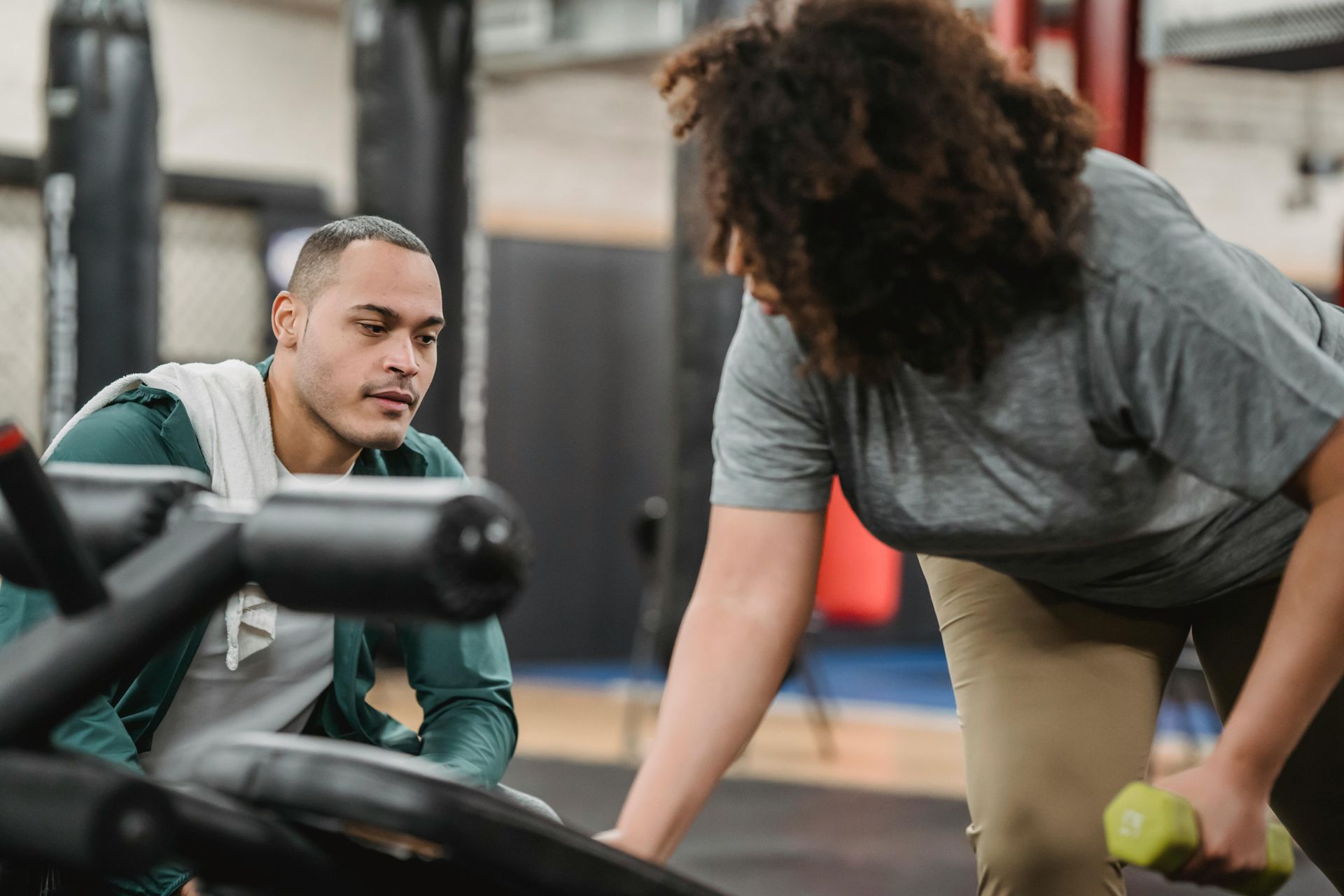 Gym trainer assisting a person lifting a dumbbell. Inside a gym with equipment, one person is bent over.