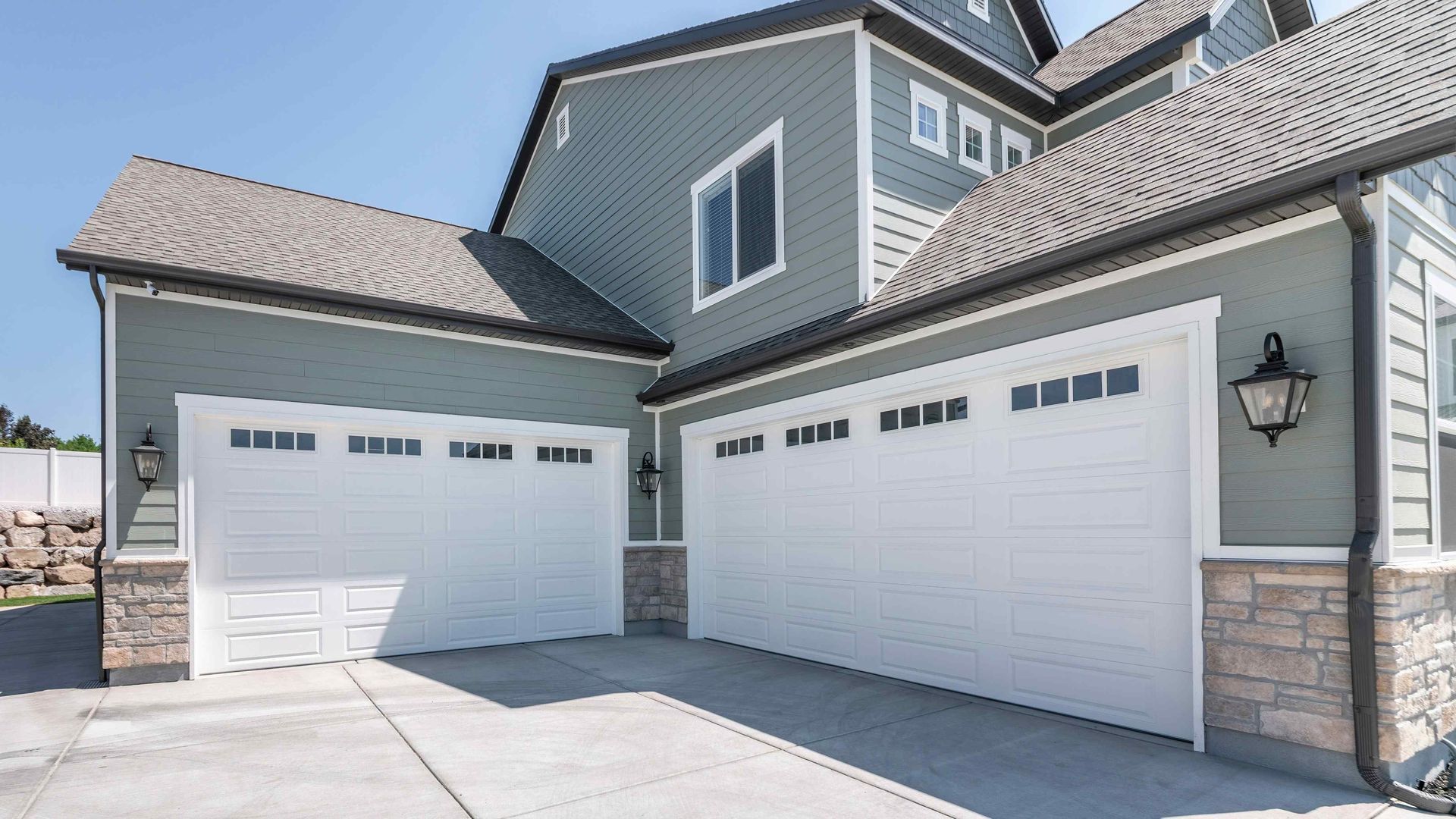 A two-story suburban home with sage green siding, stone accents, and two white garage doors under a clear blue sky.