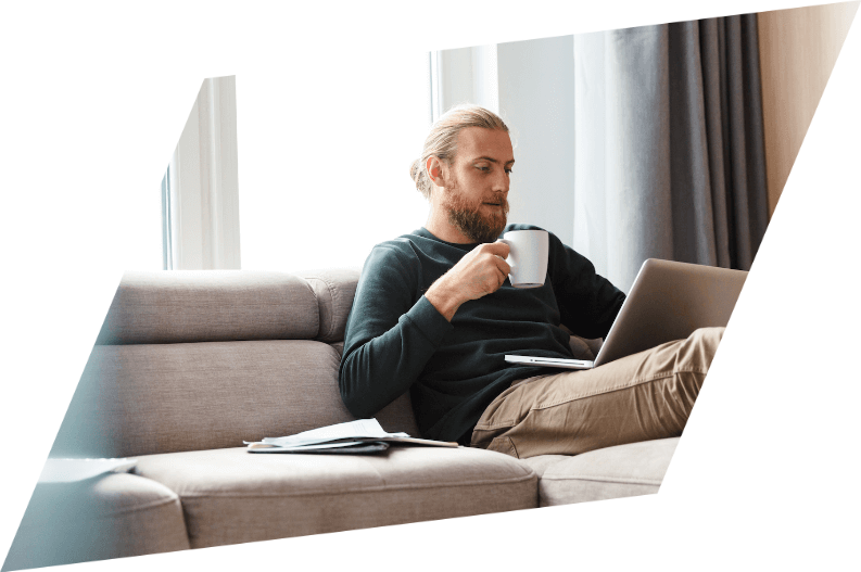 A man is sitting on a couch drinking coffee and using a laptop.