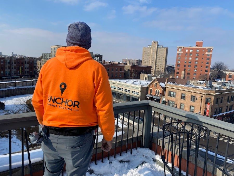 A man wearing an orange anchor sweatshirt is standing on a balcony overlooking a snowy city.