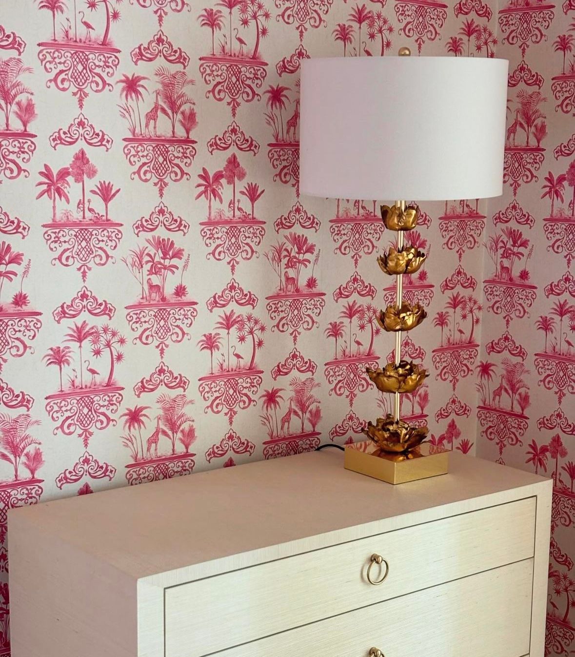 A lamp sits on a dresser in front of a pink and white wallpaper