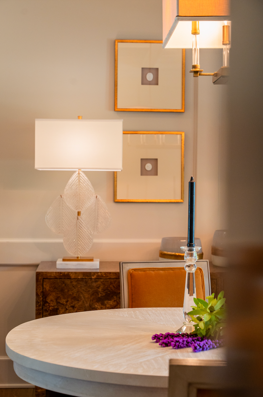 A dining room with a table , chairs , lamps , and paintings on the wall.