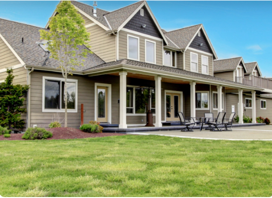 Large, two-story house with beige siding, dark trim, and a wide porch. Green lawn and a clear blue sky.