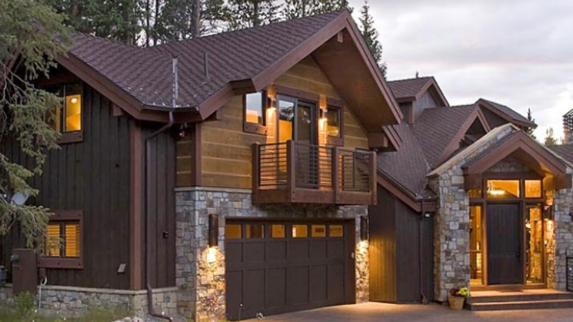 Brown and stone exterior of a two-story home with a garage, balcony, and front entrance, nestled in a wooded setting.