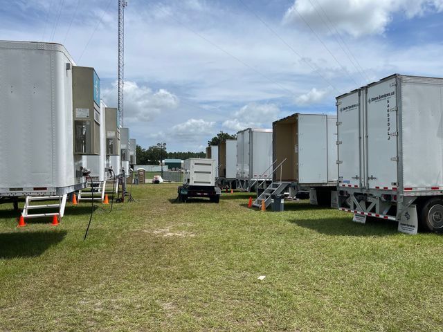 Two white trailers on a wet road, overcast sky. Trees and a structure in background.