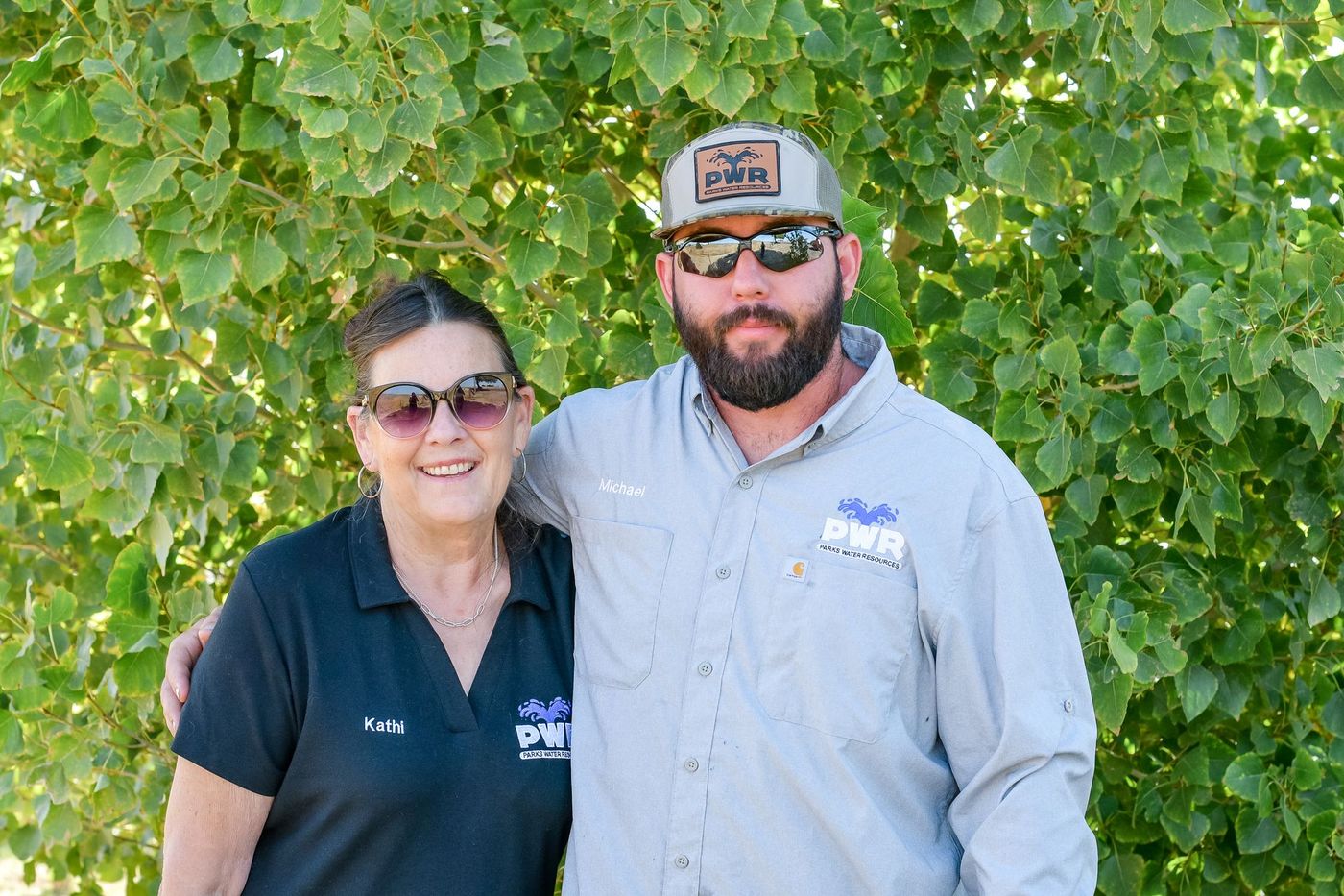 Woman and man smiling, posing outdoors, with green foliage background. They are wearing sunglasses and shirts with 
