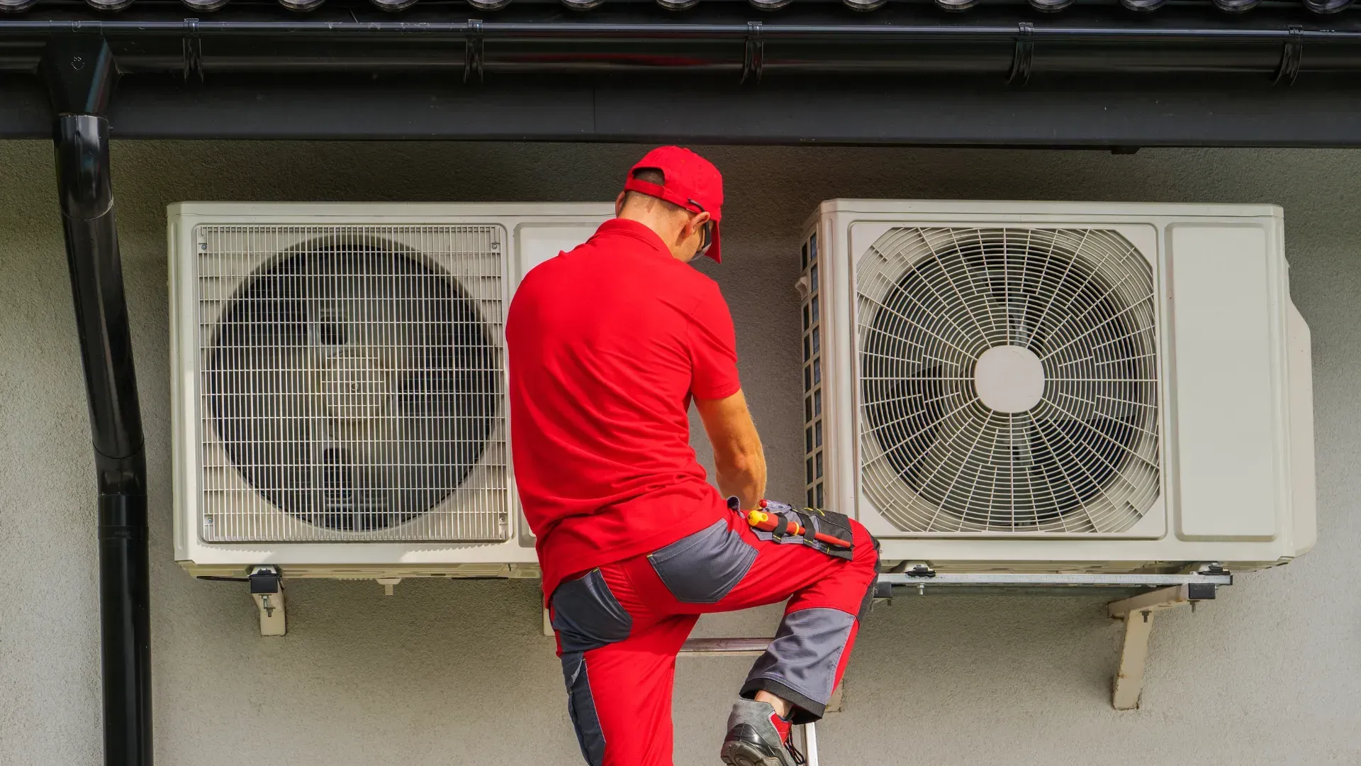 A man in a red shirt is working on an air conditioner on the side of a building.