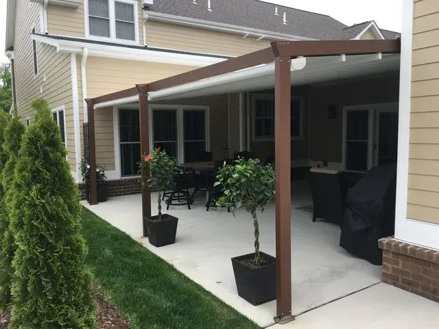 Covered patio with brown posts, black chairs, potted plants, and a grill beside a house and lawn