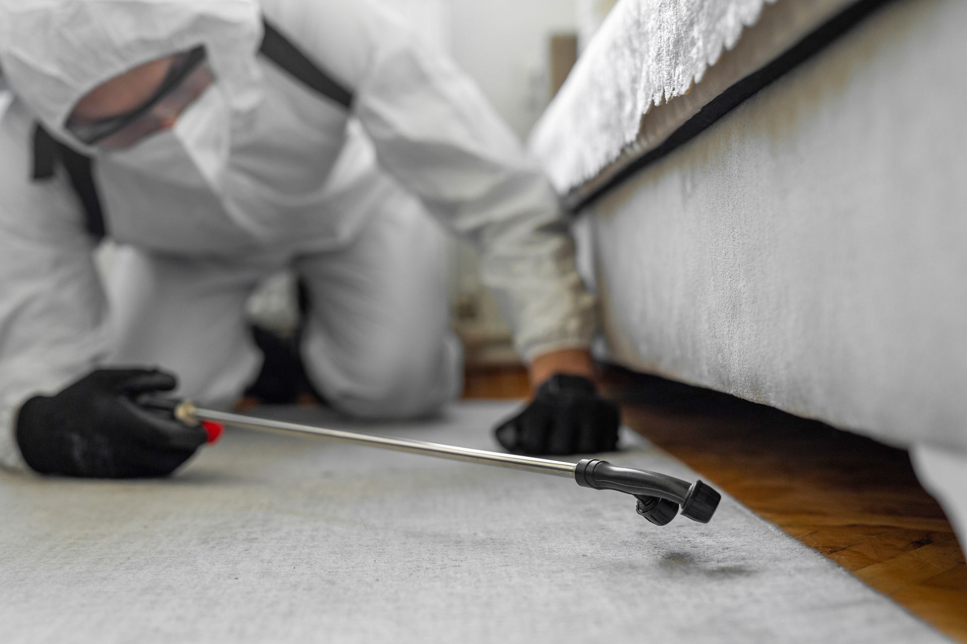 Pest control technician in a white suit sprays insecticide near a bed, presumably indoors.