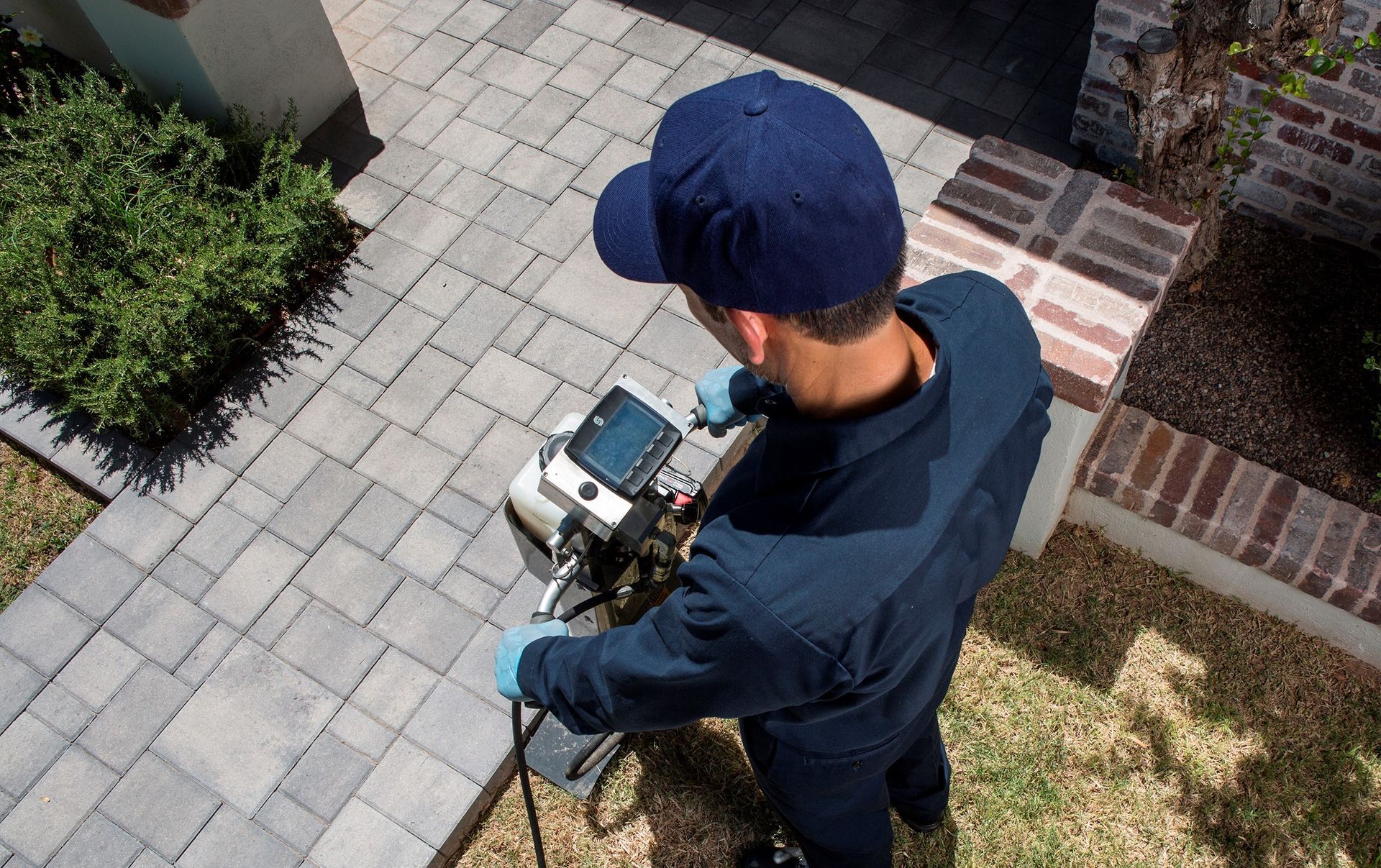 Man in blue uniform using pest control equipment on a brick patio near a house.