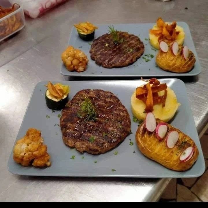 Due piatti gourmet a base di bistecca con contorni di patate e verdure su un bancone in acciaio inossidabile.