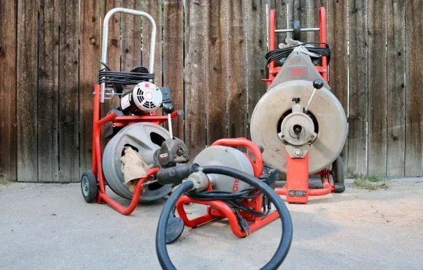 Three drain cleaners are sitting on the ground in front of a wooden fence.