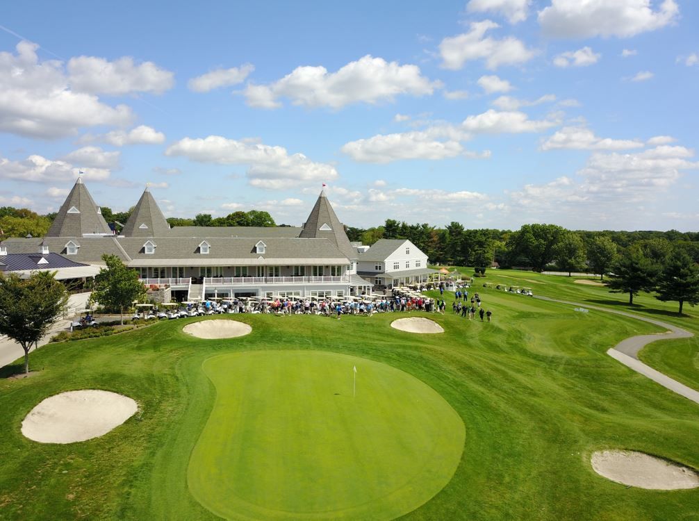 Two girls hold hands, carrying golf clubs while walking on a golf course. Family follows in the background.