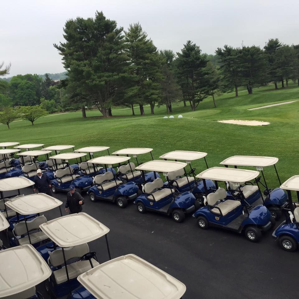 Woman in red top adjusts golf club on golf course; golfers and carts in background.