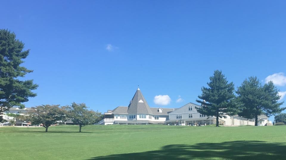 Large white building with a distinctive conical tower, set in a grassy field with trees under a blue sky.