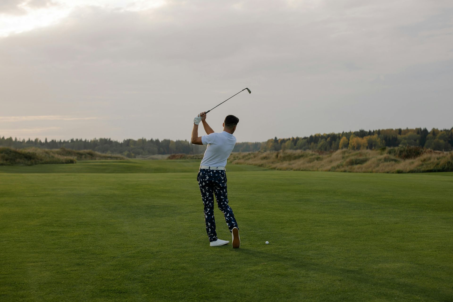 Golfer swings club on green course; cloudy sky, trees in background.