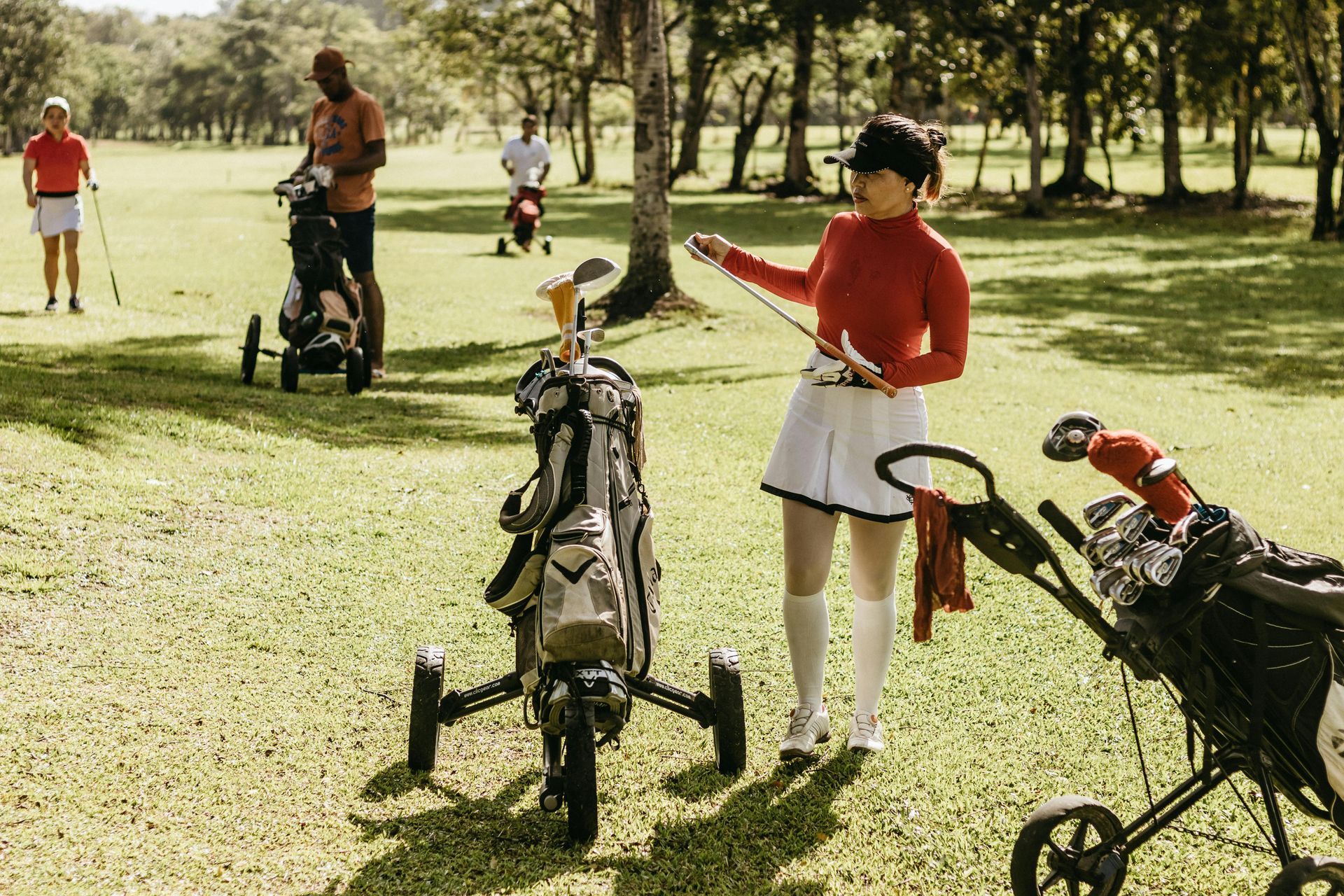 Woman in red top adjusts golf club on golf course; golfers and carts in background.