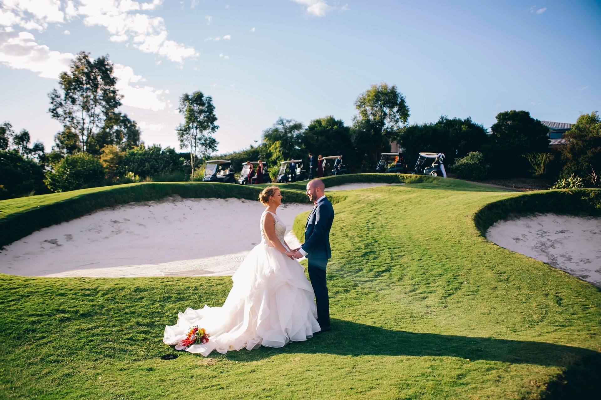 Bride and groom holding hands, standing on a golf course. She wears a white dress; he wears a navy suit. Bright, sunny day.