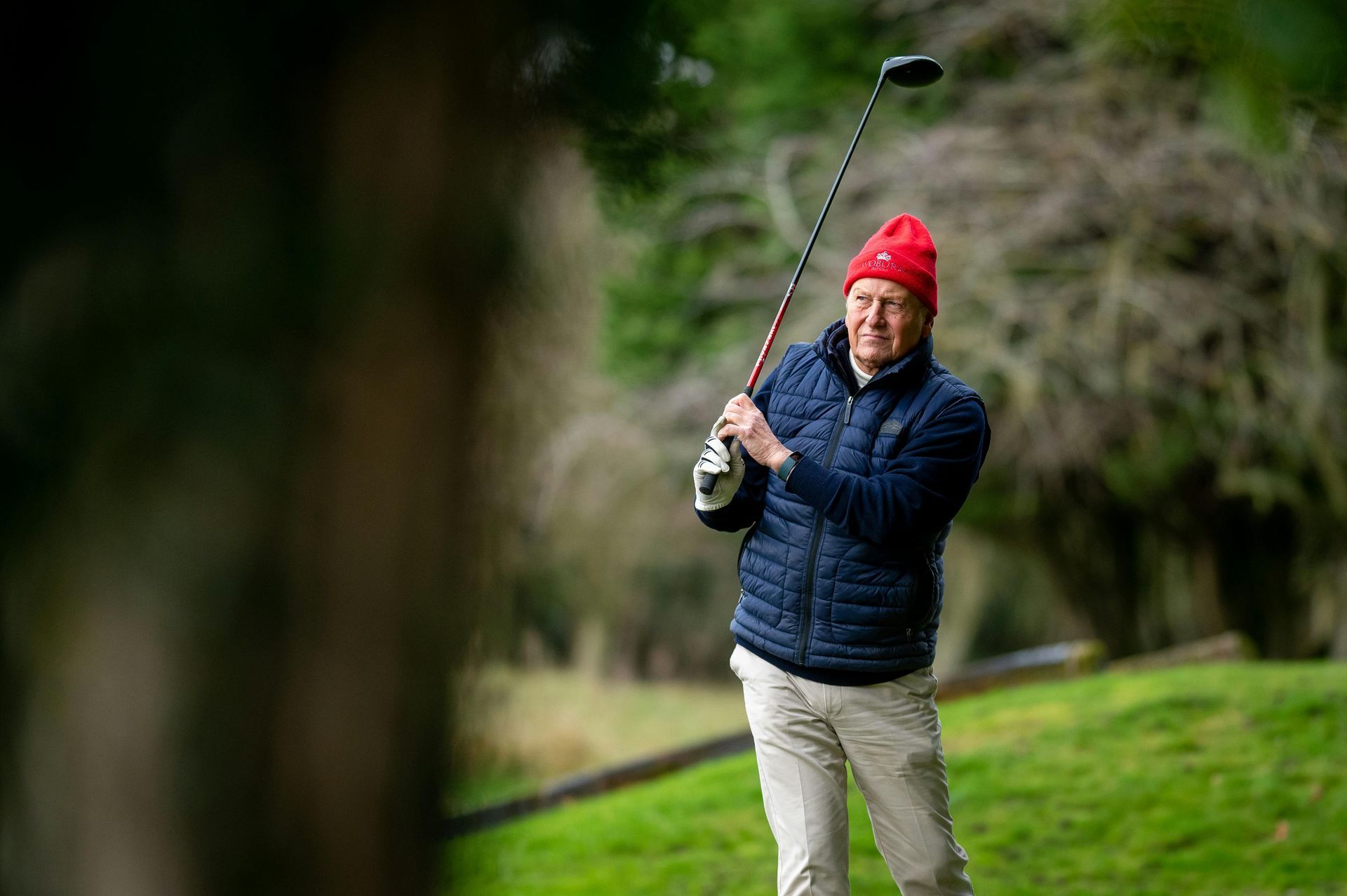 Man swings golf club on a green course, wearing a red hat and blue jacket.