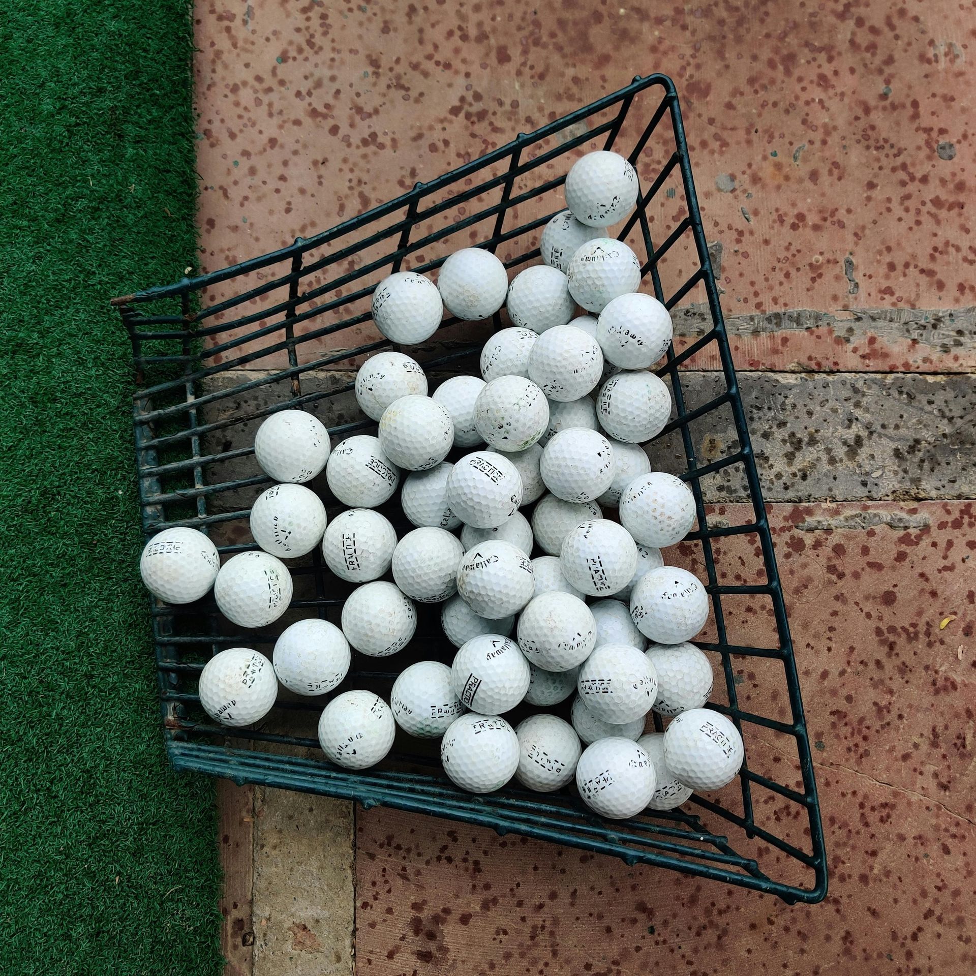 Black wire golf ball basket filled with white golf balls, placed on brick.